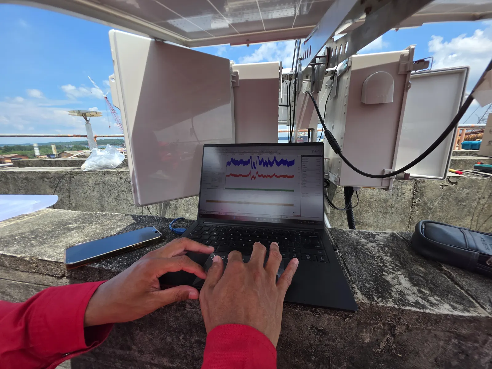 Hands typing on a laptop on a rooftop, next to open sensor enclosures, with live waveforms on screen