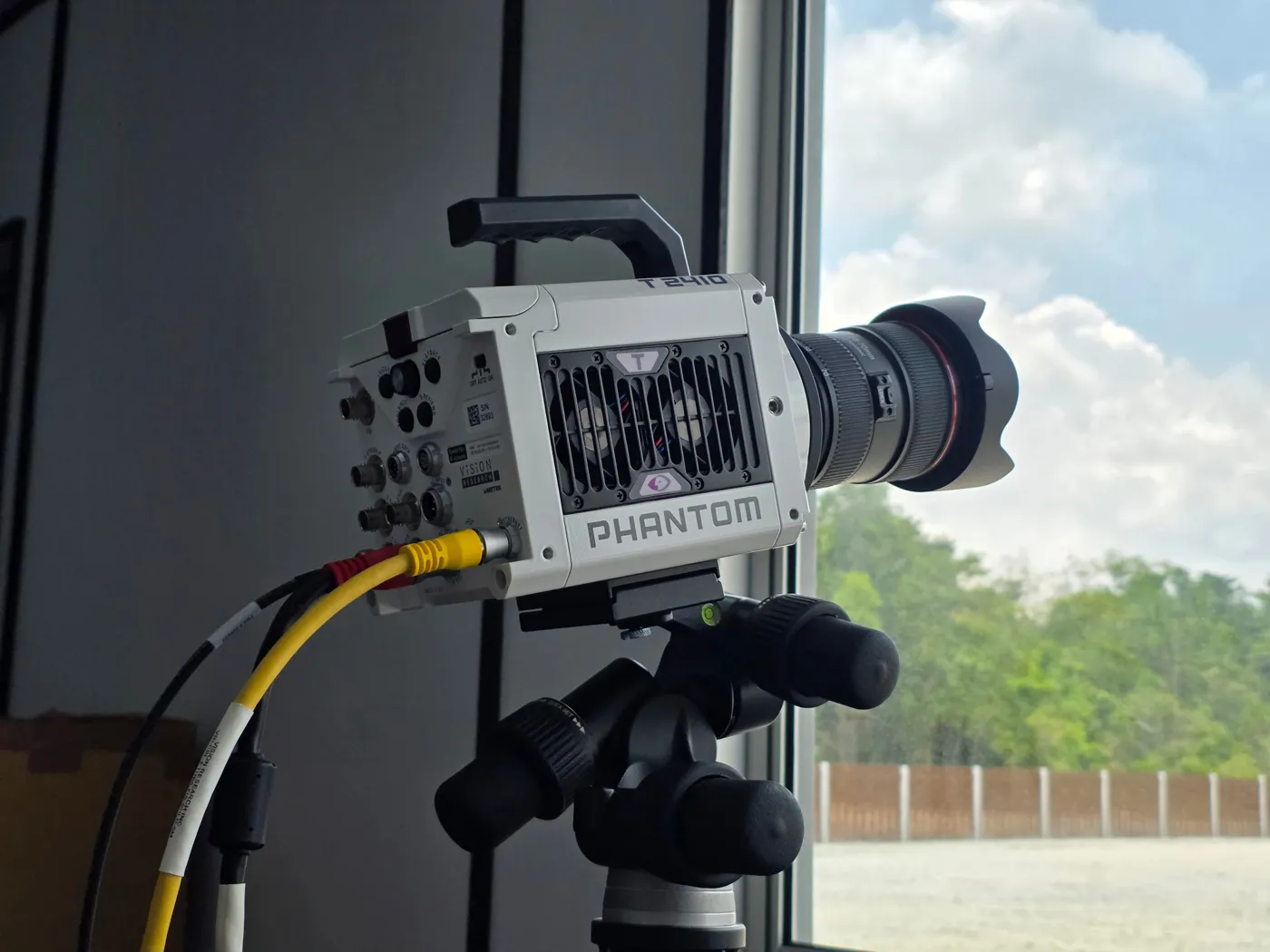 The white Phantom T2410 high-speed camera on a tripod by a window, with a zoom lens aimed at the launch field